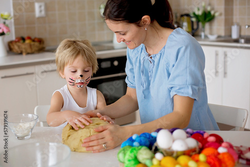 Little blond toddler child with painted face as rabbit and young mother, preparing dough for easter brioce buns, sweet easter bread
