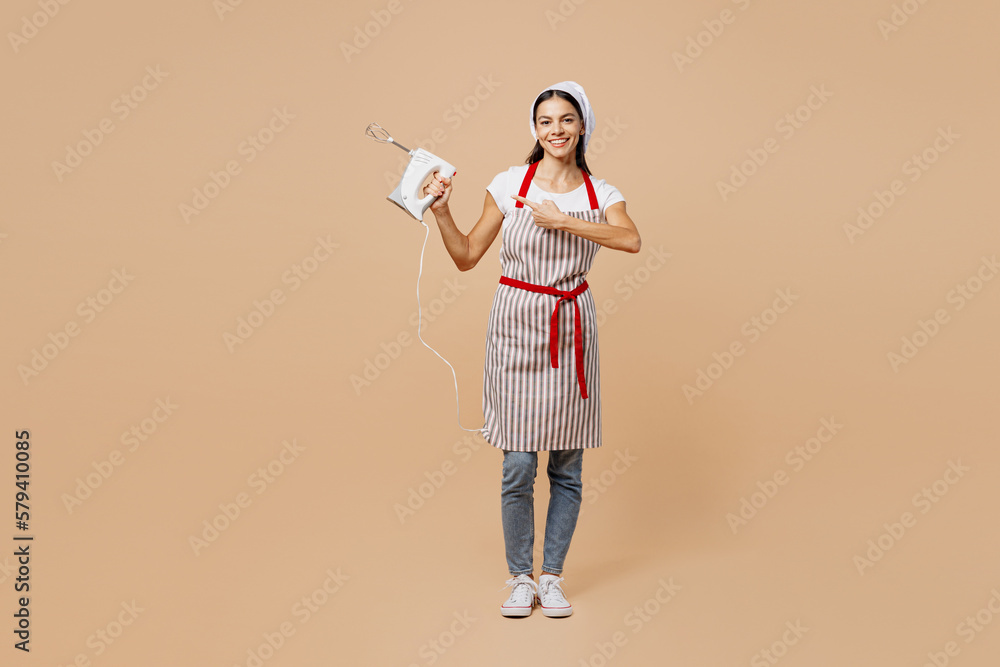 Full body young fun housewife housekeeper chef baker latin woman wear striped apron toque hat hold in hand show point finger on mixer isolated on plain pastel light beige background Cook food concept Full body young fun housewife housekeeper chef baker latin woman wear striped apron toque hat hold in hand show point finger on mixer isolated on plain pastel light beige background Cook food concept