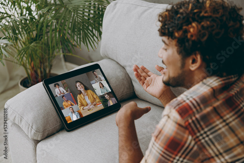 Young Indian man wears casual clothes use digital tablet pc computer leading video conference call lay down on grey sofa stay at home hotel flat rest relax spend free spare time in living room indoor.