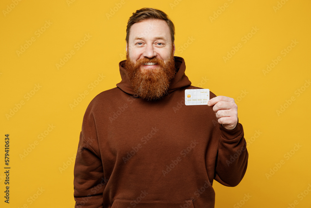 Young smiling fun redhead caucasian man wearing brown hoody casual clothes hold in hand mock up of credit bank card look camera isolated on plain yellow background studio portrait. Lifestyle concept. Young smiling fun redhead caucasian man wearing brown hoody casual clothes hold in hand mock up of credit bank card look camera isolated on plain yellow background studio portrait. Lifestyle concept.