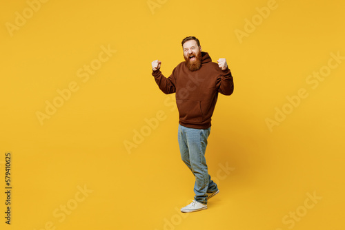 Full body young redhead caucasian man wear brown hoody casual clothes doing winner gesture celebrate clenching fists say yes walk isolated on plain yellow background studio portrait Lifestyle concept