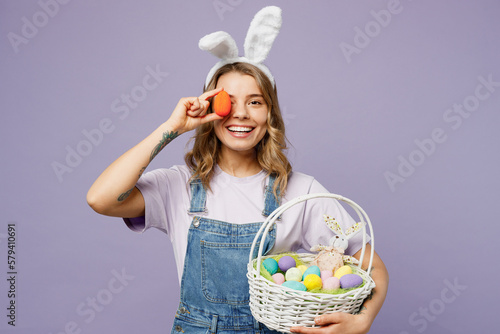 Photography Young smiling fun woman wearing casual clothes bunny rabbit ears hold wicker basket cover eye with colorful violet egg isolated on plain pastel purple background studio portrait