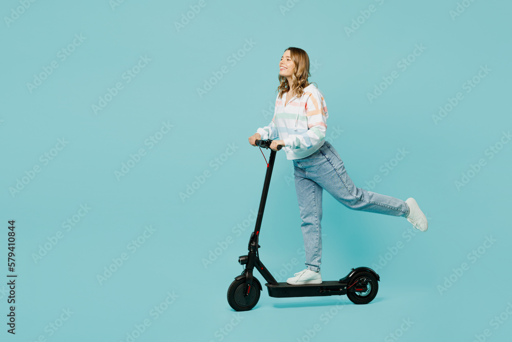 Full body side view smiling happy cheerful young woman wear striped hoody riding modern e-scooter raise up leg isolated on plain pastel light blue cyan background studio portrait. Lifestyle concept. Full body side view smiling happy cheerful young woman wear striped hoody riding modern e-scooter raise up leg isolated on plain pastel light blue cyan background studio portrait. Lifestyle concept.