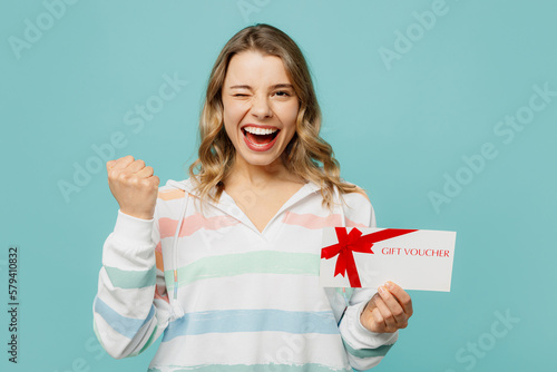 Young woman wears striped hoody hold gift certificate coupon voucher card for store do winner gesture wink blink eye isolated on plain pastel blue cyan background studio portrait. Lifestyle concept.