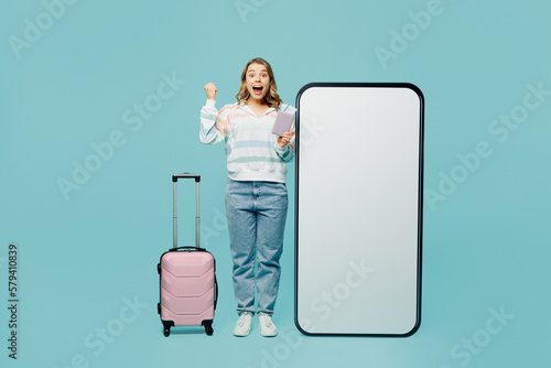 Traveler woman wear casual clothes hold suitcase passport ticket big huge blank screen mobile cell phone isolated on plain blue cyan background. Tourist travel abroad in free spare time rest getaway.