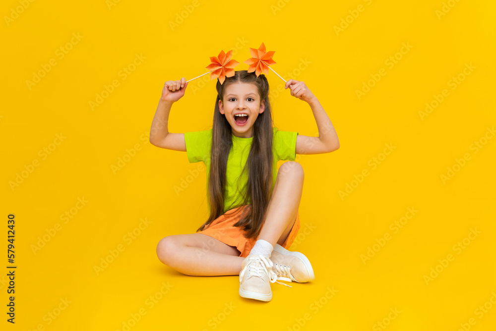 A little girl is playing with paper flowers on sticks. An origami mill. Happy kid holds paper crafts. Children's creativity. A beautiful child is sitting on a yellow isolated background. A little girl is playing with paper flowers on sticks. An origami mill. Happy kid holds paper crafts. Children's creativity. A beautiful child is sitting on a yellow isolated background.