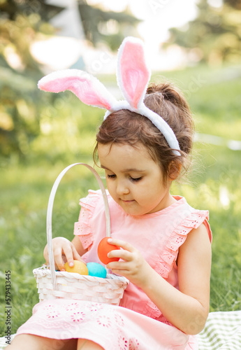 Little cute girl wear bunny ears sitting on a plaid and holding basket with colorful painted eggs on Easter egg hunt in park