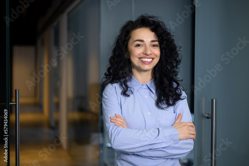 Print op canvas Portrait of happy and successful business woman, boss in blue shirt smiling and looking at camera inside office with crossed arms, Hispanic woman with curly hair in corridor
