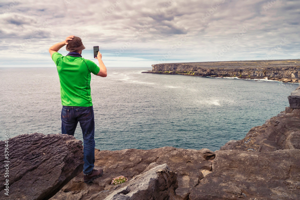 Male tourist in green shirt standing on edge of a cliff taking picture on his smart phone of stunning scenery. Aran Island, Ireland. Travel and tourism. Rough Irish nature landscape.