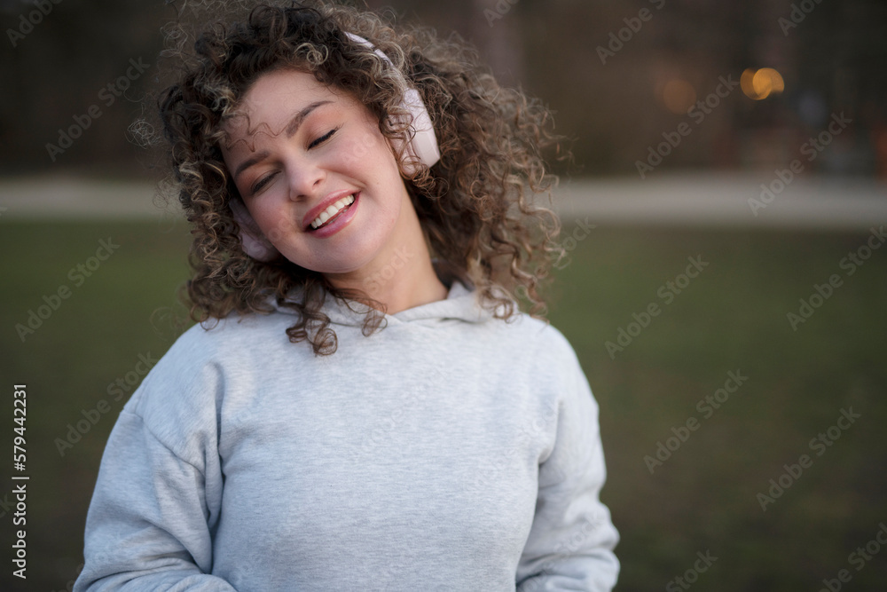 Young beautiful woman in a gray sports hoodie, wearing headphones, listening to music in the evening in the park