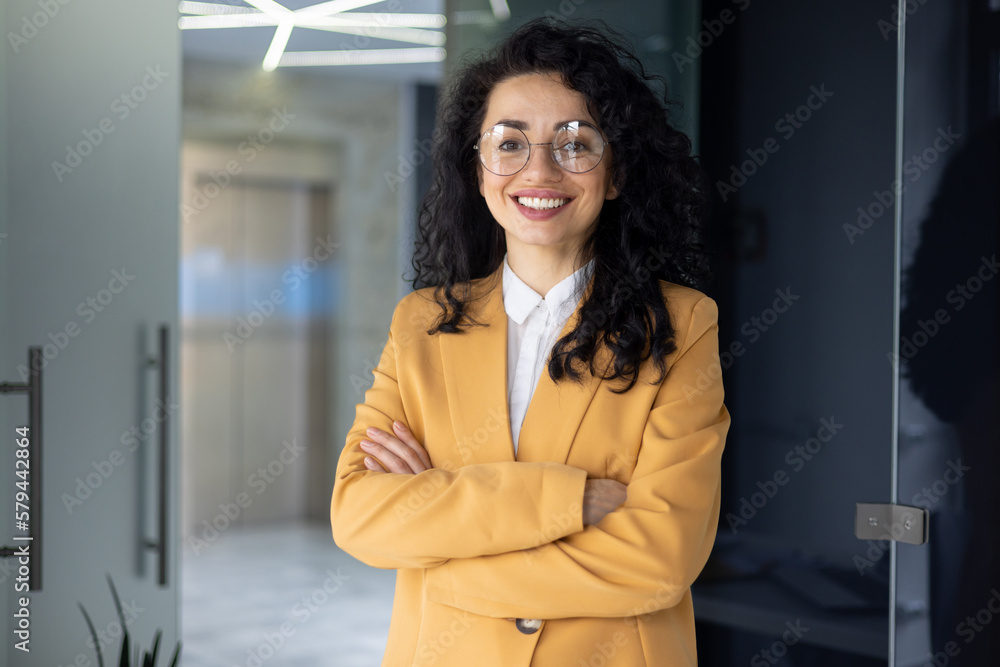 Portrait of successful businesswoman inside office, Hispanic with curly hair and glasses smiling and looking at camera, boss woman in yellow jacket, financier happy with achievement results at work. Portrait of successful businesswoman inside office, Hispanic with curly hair and glasses smiling and looking at camera, boss woman in yellow jacket, financier happy with achievement results at work.