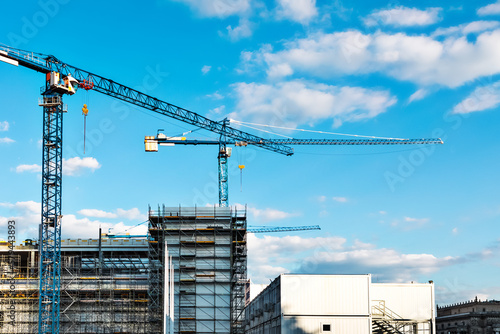 Two construction tower cranes at a construction site against a blue sky with clouds. Modern high-rise office building under construction. Copyspace top right, horizontal