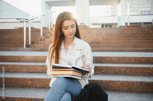 Portrait of a cheerful young girl student with backpack sitting on steps outdoors, reading book