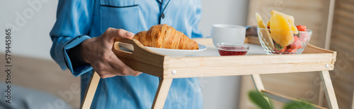 cropped view of woman in blue pajama holding tray with breakfast in bedroom, banner.