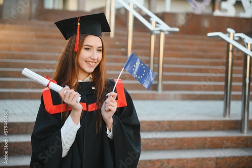Happy cute brunette caucasian grad girl is smiling. She is in a black mortar board, with red tassel, in gown, with nice brown curly hair, diploma in hand.