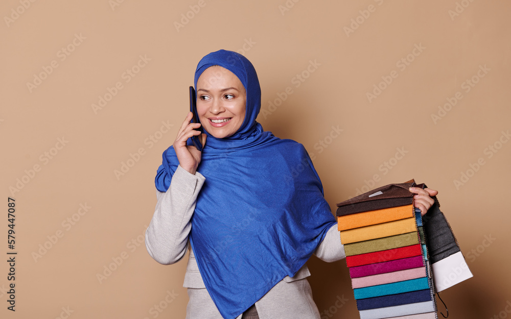 Charming Arab Muslim woman with head covered in blue hijab, confident successful sales manager, decorator talks on mobile phone, taking orders for sofa upholstery materials, isolated beige background Charming Arab Muslim woman with head covered in blue hijab, confident successful sales manager, decorator talks on mobile phone, taking orders for sofa upholstery materials, isolated beige background