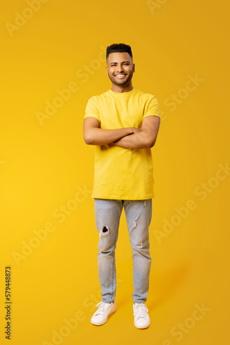 Full size length portrait of confident happy young Indian man 20s in yellow t-shirt holding hands crossed isolated on plain yellow background, foreign male student