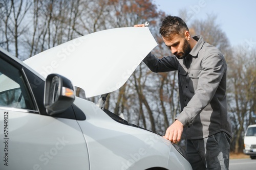 Young upset casual man trying to fix his broken car outdoors. Man waiting for towing service for help car accident on the road. Roadside assistance concept.