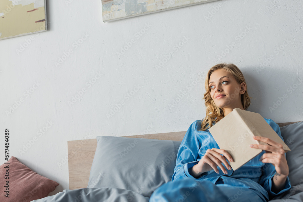 pensive young woman in blue silk nightwear holding book in bedroom. pensive young woman in blue silk nightwear holding book in bedroom.