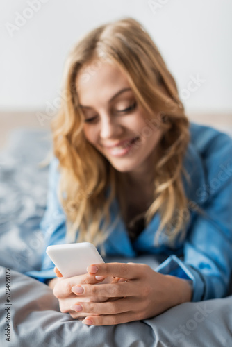 cheerful woman messaging on smartphone while lying in blue pajama on bed.