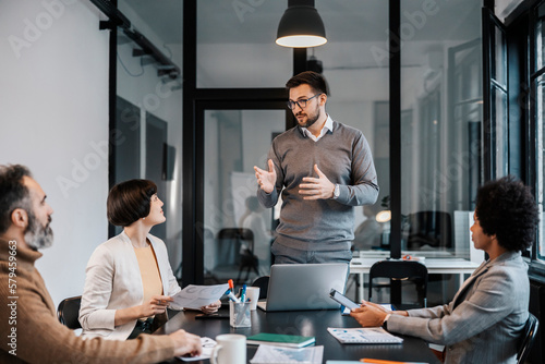 A mentor is standing at the boardroom and explaining new project while associates are sitting and listening.