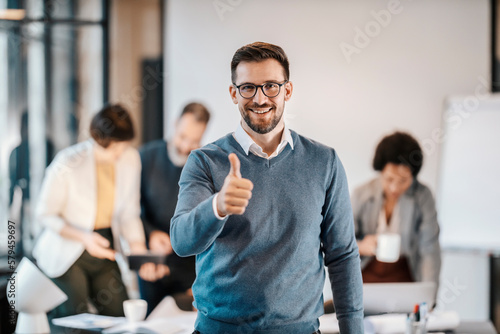 Foto Portrait of a happy businessman giving thumbs up at the camera while his colleagues working in a blurry background at the office