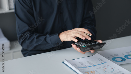 A businessman is using a calculator to calculate financial numbers, he is sitting in his private office, the businessman examines the financial data from the corporate finance chart.