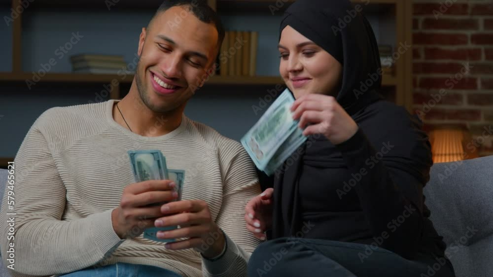 Diverse ethnic couple sit on sofa counting money dollars banknotes