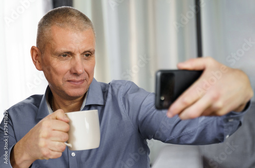 A man with tea makes a selfie photo with a mobile phone in the apartment.