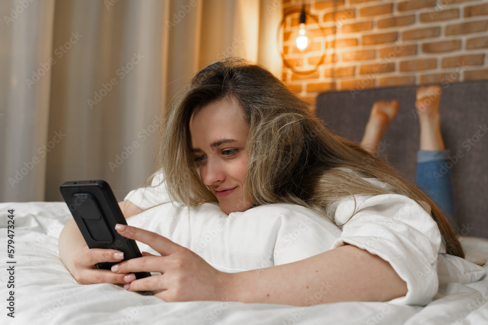 A young beautiful woman is lying on the bed, watching content in a smartphone, smiling A young beautiful woman is lying on the bed, watching content in a smartphone, smiling