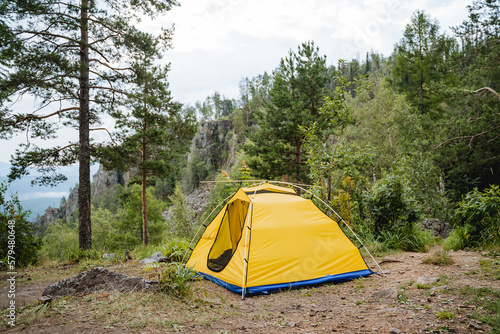Yellow tourist tent stands in a clearing in the forest, mountain climbing camping on a cliff, open entrance, no people, metal frame, hiking in the mountains