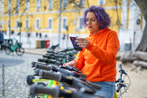 Woman Unlocking Electric Bike Through Smartphone.Mature Woman With Phone And Electric Bike Instead Of Transportation, Clean Energy Or Sustainable Travel.  Lifestyle