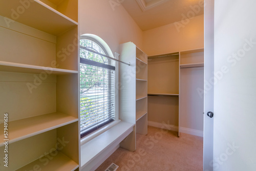 Interior view of a walk in closet of a house with built in shelves and cabinets. Interior design ideas for a new home featuring arched window and spacious storage.