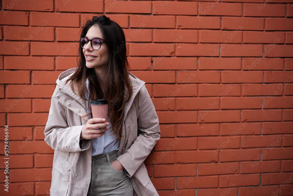 A happy smart casual woman leaning on brick wall outside and enjoying takeaway coffee. A happy smart casual woman leaning on brick wall outside and enjoying takeaway coffee.