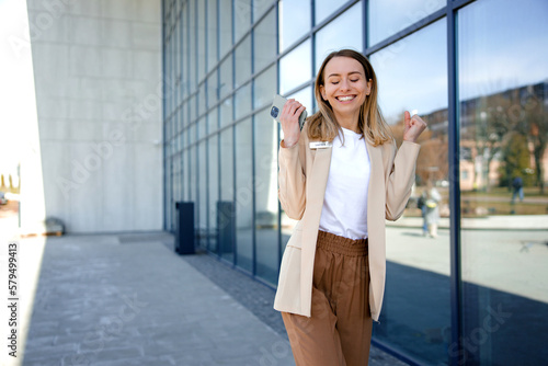 Free Download Excited caucasian happy businesswoman reading good news on smartphone while standing outdoors. Smiling lady worker in suit making yes gesture with office center on background. - GFXLoad
