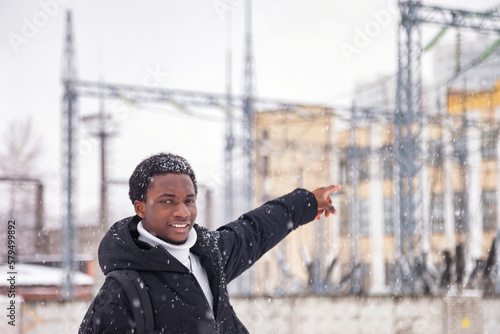 African american man pointing to side index finger in snowy winter at industry background, looking at camera. Positive black man gesturing outside, falling snowflakes. City life concept. Copy space