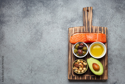 Food sources of healthy unsaturated fat and omega 3: fresh raw salmon fillet, avocado, olives, nuts on cutting board, rustic stone background top view. Healthy nutrition and keto diet, space for text