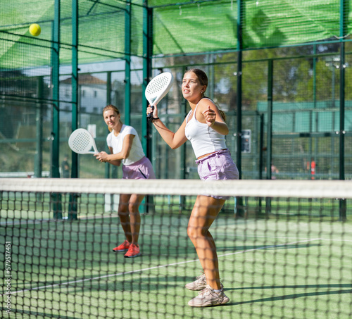 Young woman in shorts playing padel tennis on court with partner. Racket sport training outdoors.