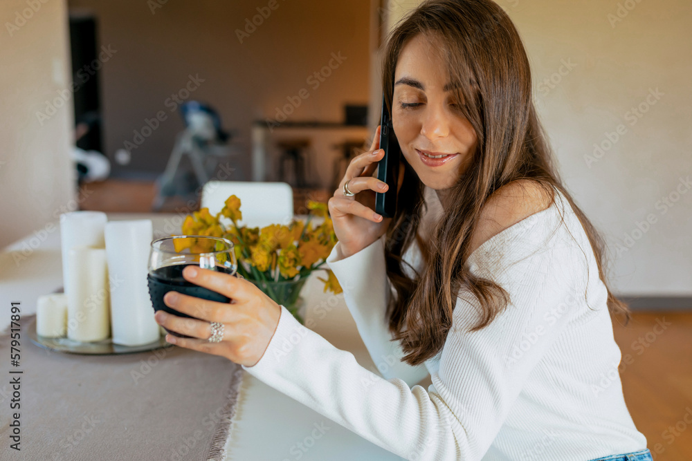 pleasing woman looking down and smiling with cup coffee . Caucasian dark hair woman in hands cup coffee, sitting at the kitchen with smartphone. Concept of lifestyle, enjoying life pleasing woman looking down and smiling with cup coffee . Caucasian dark hair woman in hands cup coffee, sitting at the kitchen with smartphone. Concept of lifestyle, enjoying life