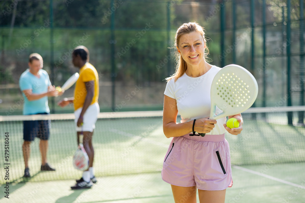 Portrait of positive caucasian woman standing on padel tennis court, holding racket and ball, smiling and looking at camera.