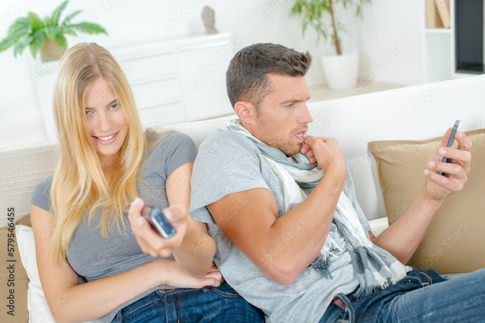 Couple on sofa, lady holding remote control, man looking at cellphone Couple on sofa, lady holding remote control, man looking at cellphone