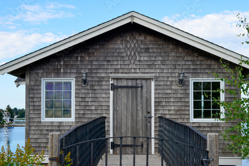 The exterior entrance to a cedar shake covered building with a grey single wooden door and two glass windows. The trim on the cottage is white. There's a black metal railing and get leading up to it.