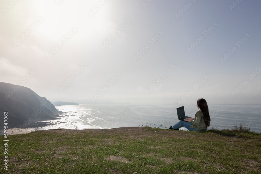 Digital nomad lifestyle concept with adult woman sit down on the ground with scenic view of beach and ocean inbackground - work with technology internet laptop computer in alternative free office