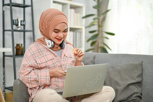 Happy young Asian Muslim woman eating cookies and watching movie on her laptop in living room.