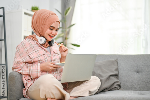 Happy young Asian Muslim woman eating cookies and watching movie on her laptop