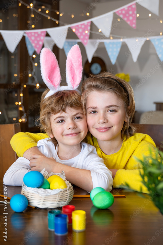 Obraz premium Easter Family traditions. Two caucasian happy children with bunny ears dye and decorate eggs with paints for holidays while sitting together at home table. Kids embrace and smile in cozy. Vertical