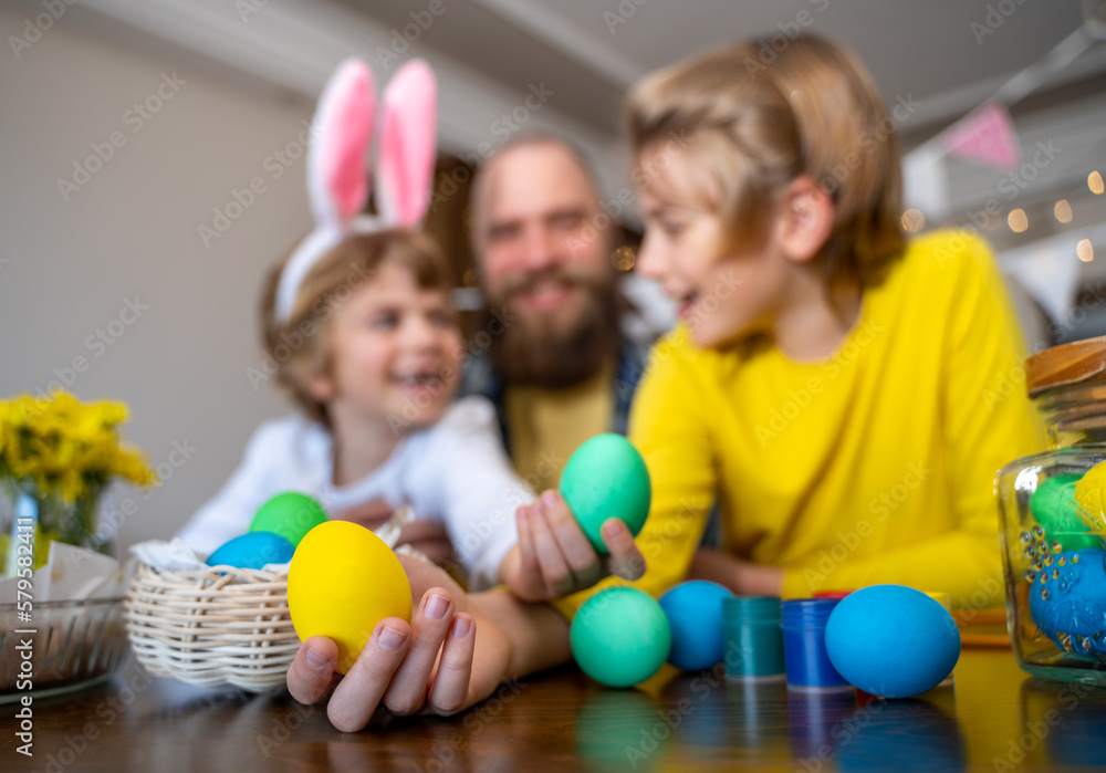 Fototapeta premium Easter Family traditions. Father and two caucasian happy children with bunny ears dye and decorate eggs with paints for holidays while sitting together at home table. Kids embrace and smile in cozy