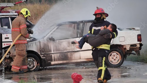 Asian fireman wear fire protection suit with a Saved boy in His Arms and Firefighter fighting with fire on a car during fire drill and helping fire victims Concept. Slow motion