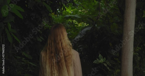 Blonde woman walking towards forest of foliage at twilight. Gimbal