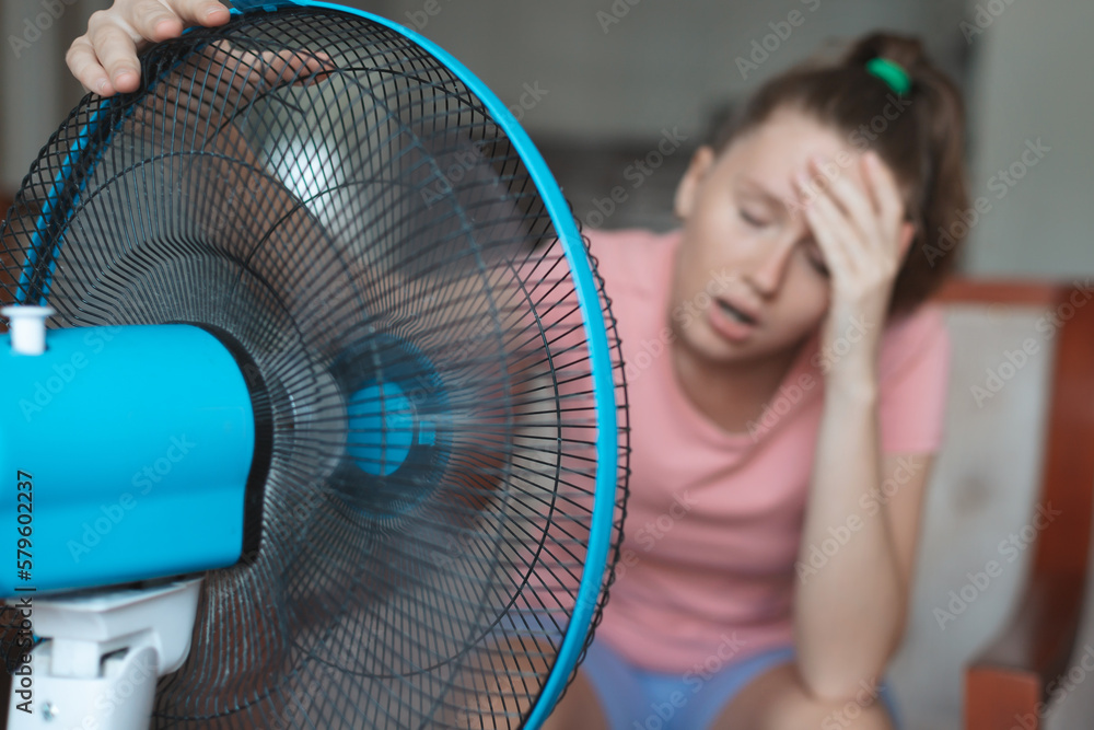 Young woman using electric fan at home in living room, sitting on couch